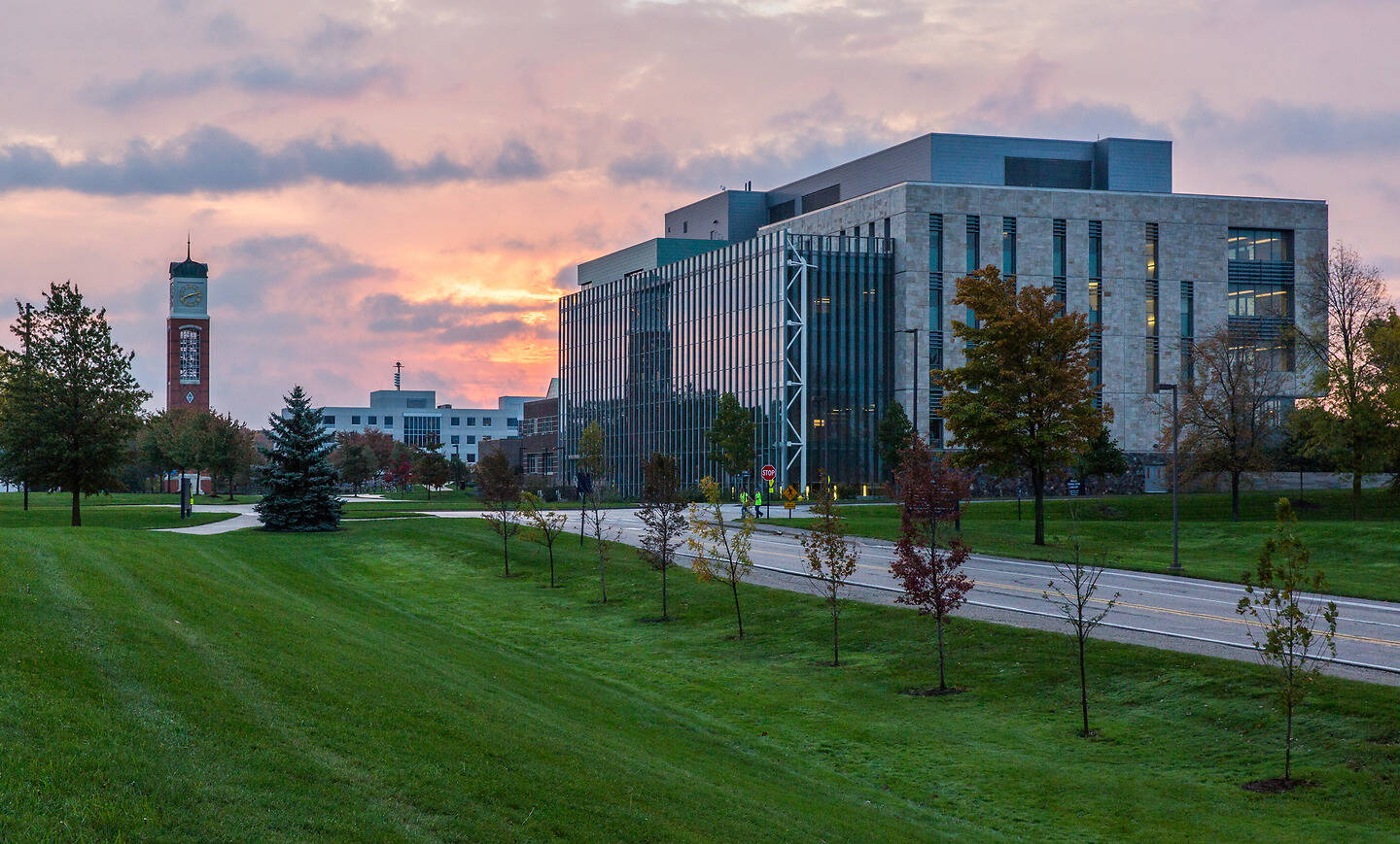 A modern building with glass and stone stands beside a grassy field under a pink and orange sunset, with a clock tower visible in the distance.